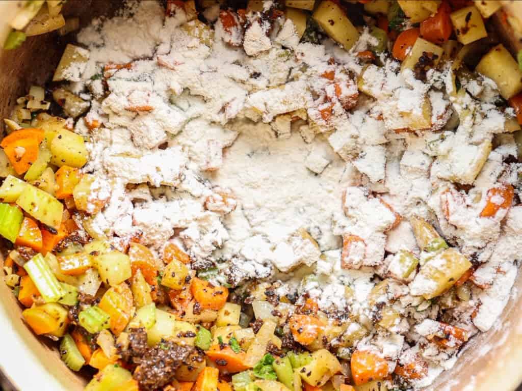 Diced carrots, celery, and potatoes are sautéed in a pot with visible specks of flour sprinkled on top, preparing the base for a savory dish.